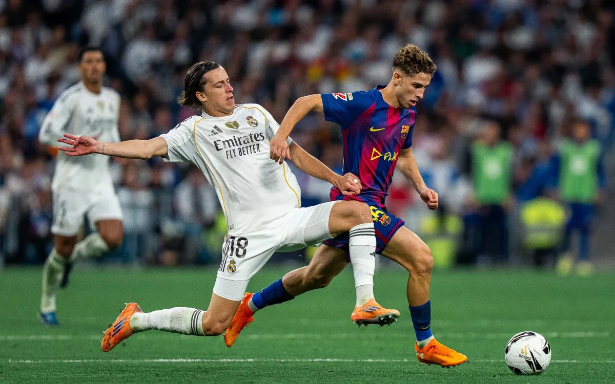 Two soccer players compete for a ball on a green field. One wears a white kit and the other a red and blue striped kit. The stadium is packed.