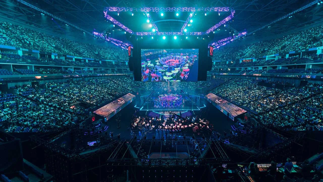 Packed stadium with a central stage lit in blue, showcasing a large screen. Spectators fill the seats, creating an energetic atmosphere.